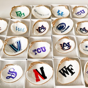 Op de afbeelding: Witte schelpen schaaltjes met goudkleurige randen, elk met een universiteitslogo in verschillende kleuren. De schelpen worden gepresenteerd in individuele witte dozen, met een verscheidenheid aan school emblemen. Een verzameling decoratieve items.