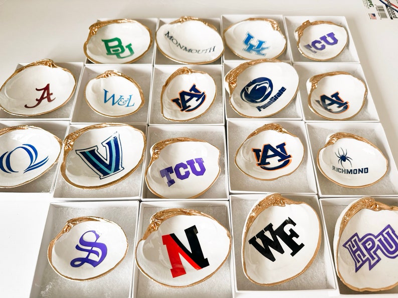 Op de afbeelding: Witte schelpen schaaltjes met goudkleurige randen, elk met een universiteitslogo in verschillende kleuren. De schelpen worden gepresenteerd in individuele witte dozen, met een verscheidenheid aan school emblemen. Een verzameling decoratieve items.