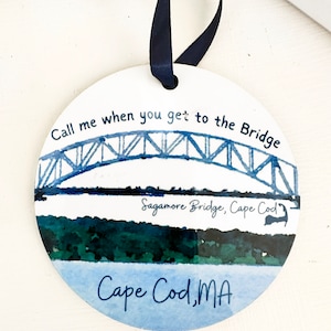 May include: Round white ornament with a watercolor painting of the Sagamore Bridge in Cape Cod, Massachusetts. The text "Call me when you get to the Bridge" is written above the bridge. The text "Sagamore Bridge, Cape Cod" is written below the bridge. The text "Cape Cod, MA" is written below the bridge.