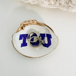 Op de afbeelding: Een witte oesterschelp sieradenschaal met een goudkleurige rand en de letters "TCU" in blauw. Twee zilveren ringen liggen in de schaal. De schaal staat op een wit oppervlak.