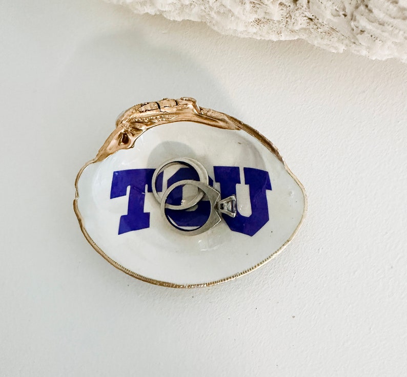 Op de afbeelding: Een witte oesterschelp sieradenschaal met een goudkleurige rand en de letters "TCU" in blauw. Twee zilveren ringen liggen in de schaal. De schaal staat op een wit oppervlak.