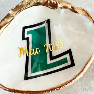 Op de afbeelding: Een witte schelpschaal met een goudkleurige rand. De schaal heeft een groene en zwarte letter "L" met het woord "Mac" en het nummer "2025" in gouden letters. De schelp ligt op een wit oppervlak.