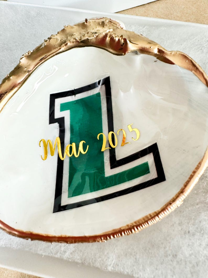 Op de afbeelding: Een witte schelpschaal met een goudkleurige rand. De schaal heeft een groene en zwarte letter "L" met het woord "Mac" en het nummer "2025" in gouden letters. De schelp ligt op een wit oppervlak.
