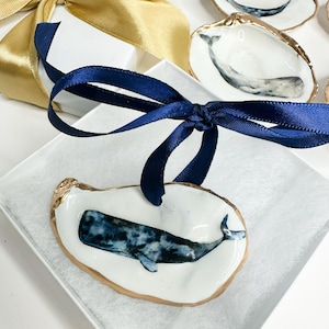 May include: A white oyster shell ornament featuring a hand-painted blue and black sperm whale, finished with a gold-painted edge and a dark blue ribbon for hanging, presented in a white gift box with a gold ribbon.