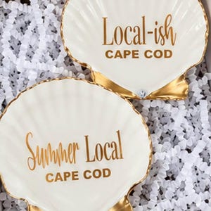 May include: Two white ceramic scallop shell dishes with gold-painted edges and bases. The top dish features the gold text Local-ish CAPE COD, while the bottom dish displays the gold text Summer Local CAPE COD in a mix of script and block lettering.