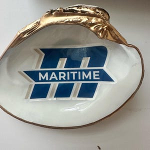 Op de afbeelding: Een decoratieve schelpschaal met een goudkleurige rand en een wit interieur. De schaal heeft een blauw en wit logo met de tekst "MARITIME". De schelp heeft een natuurlijke vorm en voegt een vleugje kustcharme toe.