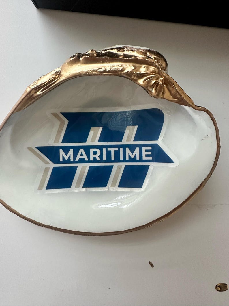 Op de afbeelding: Een decoratieve schelpschaal met een goudkleurige rand en een wit interieur. De schaal heeft een blauw en wit logo met de tekst "MARITIME". De schelp heeft een natuurlijke vorm en voegt een vleugje kustcharme toe.