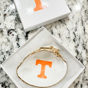 Op de afbeelding: Een witte schelpen sieradenschaal met een gouden rand en een oranje "T" aan de binnenkant, gepresenteerd in een witte geschenkverpakking met een bijpassende oranje "T" op het deksel. De schaal ligt in een witte, gewatteerde doos.