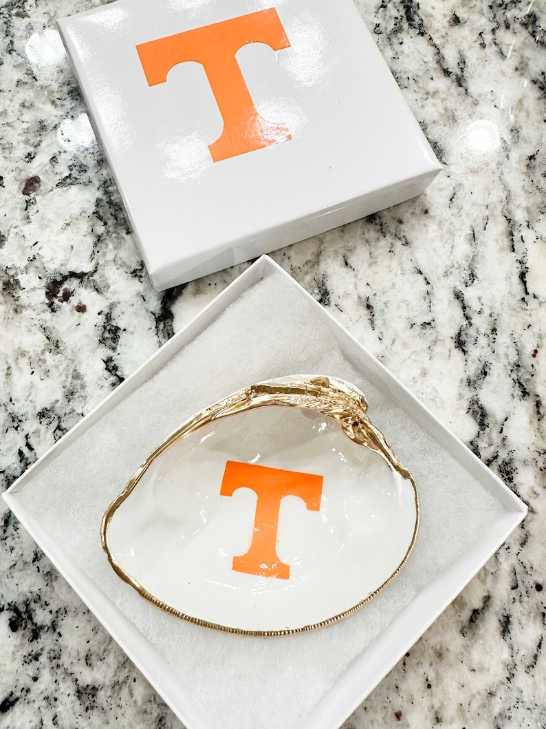 Op de afbeelding: Een witte schelpen sieradenschaal met een gouden rand en een oranje "T" aan de binnenkant, gepresenteerd in een witte geschenkverpakking met een bijpassende oranje "T" op het deksel. De schaal ligt in een witte, gewatteerde doos.