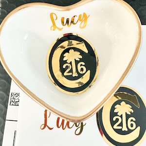 May include: A heart-shaped white ceramic dish with a gold rim, featuring the name "Lucy" in gold script. A black and gold emblem with a palm tree and the number "216" rests inside the dish. A matching emblem is next to the dish.