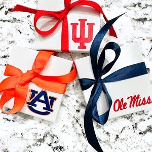 Op de afbeelding: Drie witte vierkante geschenkdozen, elk versierd met een teamlogo en een bijpassende strik. E&eacute;n doos heeft het logo van de Indiana University met een rode strik. Een andere heeft het logo van de Auburn University met een oranje strik. De derde heeft het Ole Miss-logo met een marineblauwe strik.