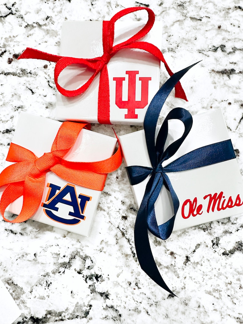Op de afbeelding: Drie witte vierkante geschenkdozen, elk versierd met een teamlogo en een bijpassende strik. E&eacute;n doos heeft het logo van de Indiana University met een rode strik. Een andere heeft het logo van de Auburn University met een oranje strik. De derde heeft het Ole Miss-logo met een marineblauwe strik.