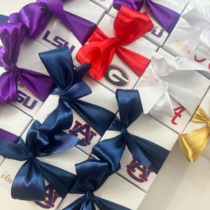 Op de afbeelding: Witte geschenkdozen met satijnen strikken in verschillende kleuren, waaronder paars, rood, marineblauw en goud. De dozen tonen logo's en tekst zoals "LSU", "AUBURN" en "Georgia". De strikken zijn bovenop de dozen geknoopt.