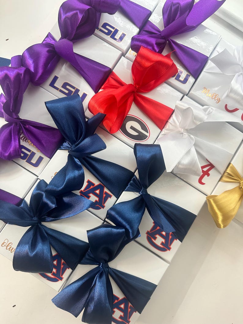 Op de afbeelding: Witte geschenkdozen met satijnen strikken in verschillende kleuren, waaronder paars, rood, marineblauw en goud. De dozen tonen logo's en tekst zoals "LSU", "AUBURN" en "Georgia". De strikken zijn bovenop de dozen geknoopt.