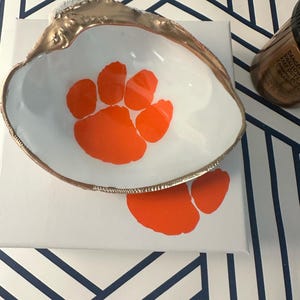 Op de afbeelding: Een wit schelpvormig schaaltje met een goudkleurige rand en een oranje pootafdrukontwerp. Het schaaltje staat op een wit oppervlak met een extra oranje pootafdruk. Een fles is zichtbaar op de achtergrond.