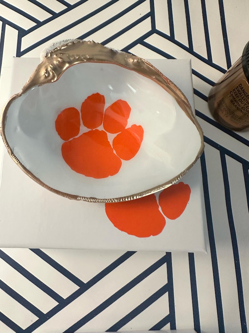 Op de afbeelding: Een wit schelpvormig schaaltje met een goudkleurige rand en een oranje pootafdrukontwerp. Het schaaltje staat op een wit oppervlak met een extra oranje pootafdruk. Een fles is zichtbaar op de achtergrond.