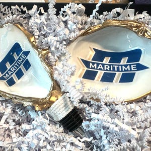 Op de afbeelding: Twee witte oesterschelpen met gouden randen, elk met een blauw en wit "MARITIME" logo. Een flesstopper met een zwarte bovenkant en witte touwdetails is ook aanwezig. De schelpen zijn genesteld in witte papieren vulling.
