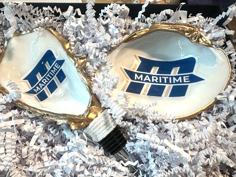 Op de afbeelding: Twee witte oesterschelpen met gouden randen, elk met een blauw en wit "MARITIME" logo. Een flesstopper met een zwarte bovenkant en witte touwdetails is ook aanwezig. De schelpen zijn genesteld in witte papieren vulling.