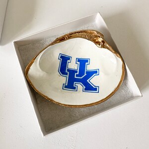 Op de afbeelding: Een wit schelpen sieradenschaaltje met een goudkleurige rand en het blauw-witte "UK"-logo. Het schaaltje wordt gepresenteerd in een witte geschenkverpakking, ideaal voor het bewaren van sieraden of kleine items.