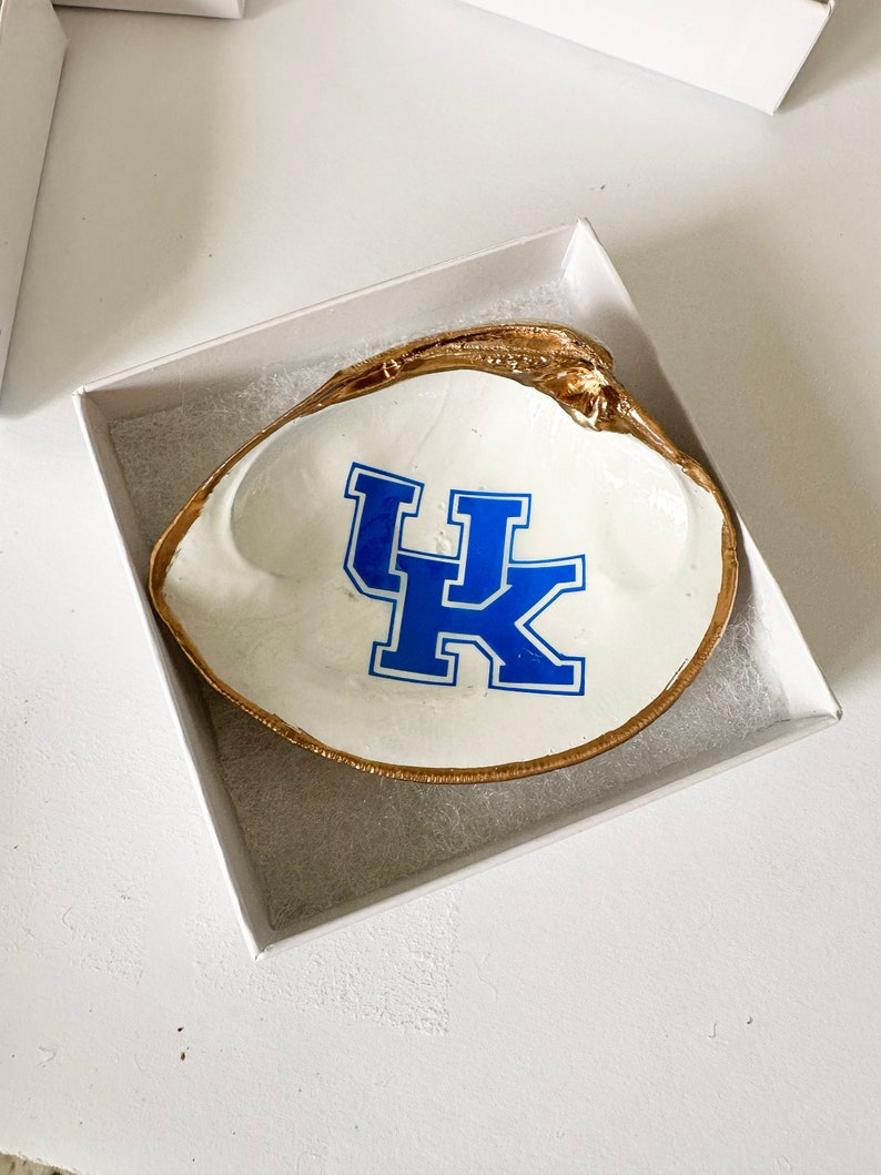 Op de afbeelding: Een wit schelpen sieradenschaaltje met een goudkleurige rand en het blauw-witte "UK"-logo. Het schaaltje wordt gepresenteerd in een witte geschenkverpakking, ideaal voor het bewaren van sieraden of kleine items.