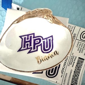 Op de afbeelding: Een wit schelpvormig sieradenschaaltje met een goudkleurige rand. Het schaaltje heeft de letters "HPU" in paars en de naam "Bianca" in goud. Het geheel ligt op een lichtblauwe ondergrond.