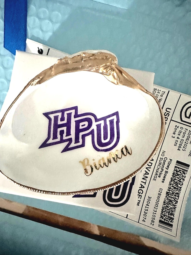 Op de afbeelding: Een wit schelpvormig sieradenschaaltje met een goudkleurige rand. Het schaaltje heeft de letters "HPU" in paars en de naam "Bianca" in goud. Het geheel ligt op een lichtblauwe ondergrond.