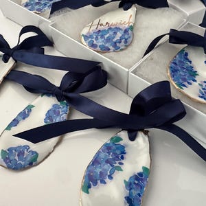May include: A collection of oyster shell ornaments featuring hand-painted blue and purple hydrangea floral patterns. Each shell has a gold-painted edge and is tied with a dark navy blue satin ribbon. One shell displays the handwritten text Harwich.