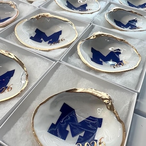 Op de afbeelding: Witte schelpvormige sieradenschaaltjes met een gouden rand, marineblauw "M"-logo en tekst "2026". De schaaltjes worden gepresenteerd in individuele witte dozen. De schelpen hebben een gemarmerd effect.