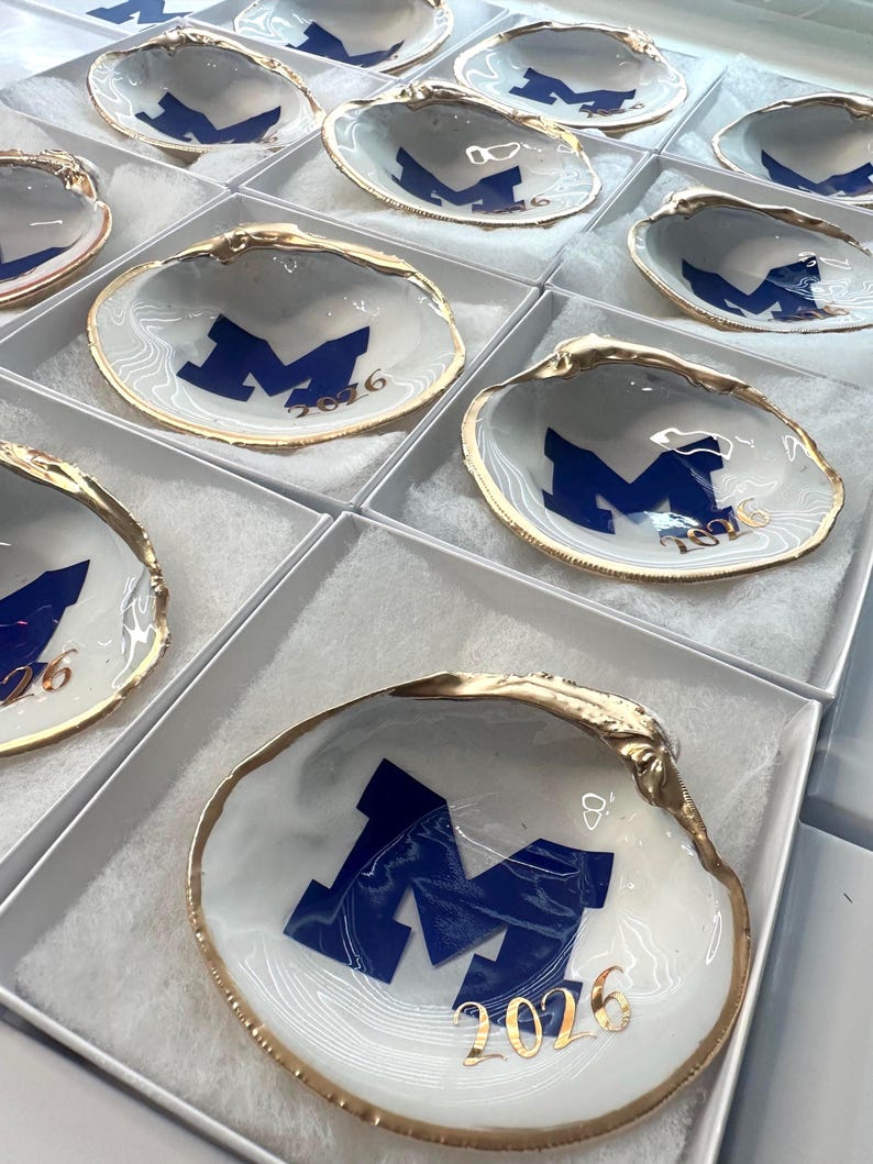 Op de afbeelding: Witte schelpvormige sieradenschaaltjes met een gouden rand, marineblauw "M"-logo en tekst "2026". De schaaltjes worden gepresenteerd in individuele witte dozen. De schelpen hebben een gemarmerd effect.
