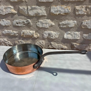Puede incluir: Una sartén sauté redonda de cobre con un interior de acero inoxidable pulido y un mango largo de metal oscuro sujeto con tres remaches. El exterior de la sartén presenta un acabado de cobre metálico cálido con un sello de fabricante visible.