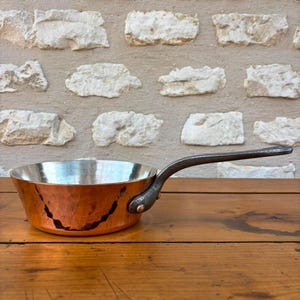 May include: A round, hammered copper sauté pan with a polished silver-toned interior lining. The pan features a long, dark, curved metal handle securely attached to the side with a single visible rivet. The exterior surface shows a textured finish.