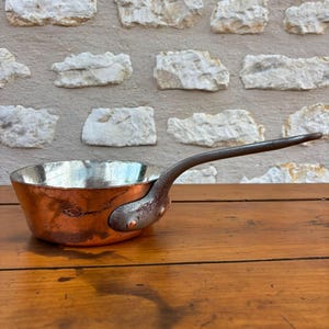 Peut inclure: Une casserole ronde en cuivre avec un revêtement intérieur argenté. L'ustensile est doté d'une longue poignée métallique incurvée de couleur sombre, fixée sur le côté par deux rivets visibles. La surface extérieure en cuivre présente une patine.