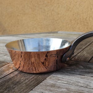 May include: A copper saucepan with a hammered texture and a dark metal handle. The interior is a shiny silver colour. The pan is on a weathered wooden surface.