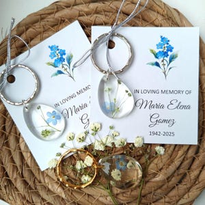 Può includere: Portachiavi a goccia in resina trasparente con fiori blu e bianchi, attaccati ad anelli portachiavi argentati. Ogni portachiavi è abbinato a un biglietto con la scritta "IN LOVING MEMORY OF Maria Elena Gomez 1942-2025" e un motivo floreale ad acquerello.