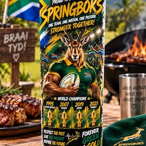 Op de afbeelding: Een cilindervormige beker met een afbeelding van een Springboks-rugbyspeler met een bal, de Zuid-Afrikaanse vlag en vier gouden kampioenschapstrofeeën met de jaartallen 1995, 2007, 2019 en 2023. Tekst bevat Proud to Support the Springboks, Stronger Together en Forever Green & Gold.