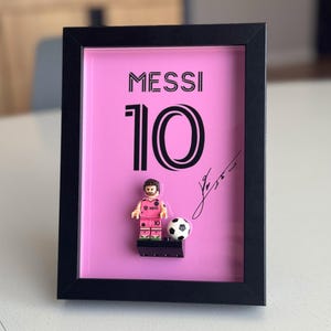 Może przedstawiać: Wzorowany druk w kolorze różowym i czarnym w ramie z figurką Lego piłkarza w różowej koszulce z numerem 10 i napisem "MESSI 10" w kolorze czarnym. Figurka trzyma piłkę nożną i stoi na czarnej podstawie. Druk zawiera również czarny podpis.
