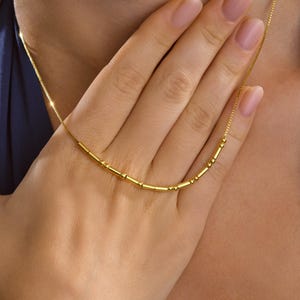 Peut inclure: Un collier en or délicat avec une série de petites perles cylindriques et de minuscules sphères. Le collier est présenté sur un fond doux, soulignant son design élégant. La chaîne est fine et l'esthétique générale est minimaliste.