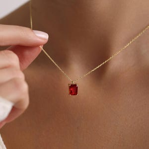 May include: A gold necklace featuring a rectangular, red gemstone pendant. The pendant hangs from a fine chain. The necklace is shown against a skin-toned backdrop.
