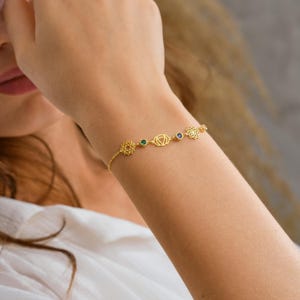 May include: A gold-colored bracelet featuring chakra symbols and small gemstones. The bracelet is worn on a wrist, showcasing its delicate design and intricate details. The bracelet's design includes geometric shapes and small, colorful accents.