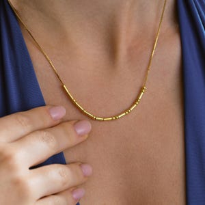 May include: A delicate gold necklace with a series of small, cylindrical and spherical beads. The necklace is worn against a blue garment. The chain is thin and the beads are evenly spaced, creating a minimalist design.