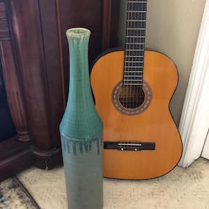 May include: A tall, cylindrical ceramic vase with a two-tone green glaze. The top portion is a vibrant teal, transitioning to a darker, dripped effect, and fading into a muted gray-green. A classical acoustic guitar leans against the wall in the background.