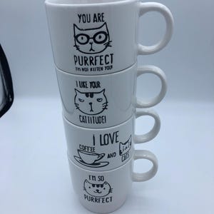 May include: Four white ceramic mugs stacked, each with a different black cat illustration and text. The mugs have phrases such as "YOU ARE PURRFECT" and "I LOVE COFFEE AND CATS". Each mug has a curved handle.