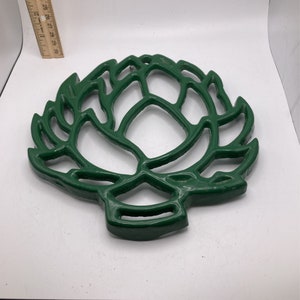 Puede incluir: Un salvamanteles de hierro fundido verde con forma de alcachofa. El salvamanteles está diseñado para proteger las superficies de los platos calientes.