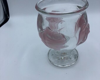 May include: Clear glass vase with a rounded body and a pedestal base. The vase features pink rose designs on the exterior. The rim of the vase is smooth and rounded. The vase is empty.