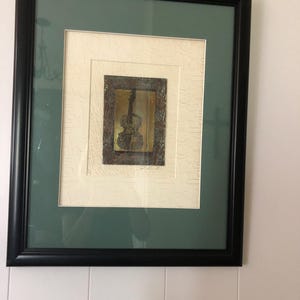 May include: A framed print of a gold-toned violin with a textured background. The frame is black with a green mount.