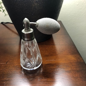 Puede incluir: Un frasco de perfume vintage con una base de vidrio transparente y facetado y un atomizador plateado. El atomizador tiene un diseño bulboso y texturizado. El frasco está sobre una superficie de madera oscura, con un jarrón oscuro en el fondo.