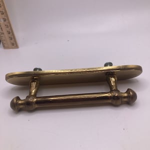 May include: A brass-coloured cabinet pull with a rectangular backplate and a cylindrical handle. Two screws secure the pull. The handle has decorative, rounded ends. Suitable for cabinets and drawers. The pull measures approximately 15 cm in length.