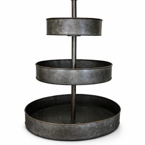以下が含まれることがあります： 素朴な亜鉛メッキ仕上げの3段メタル製トレイ。トレイは中央の垂直サポートと、さまざまなサイズの3つの円形棚を備えています。金属はまだらで古びた外観で、ヴィンテージまたはファームハウスの美学を示唆しています。