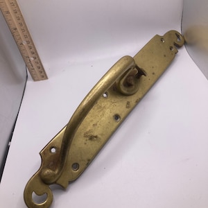May include: A brass door handle with a curved design and a decorative plate. The handle is attached to the plate with screws.