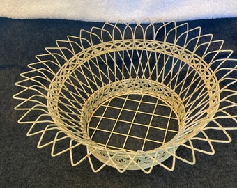 May include: A white wire basket with a circular base and a decorative, scalloped rim. The basket is made of thin wire and has a delicate, airy appearance.